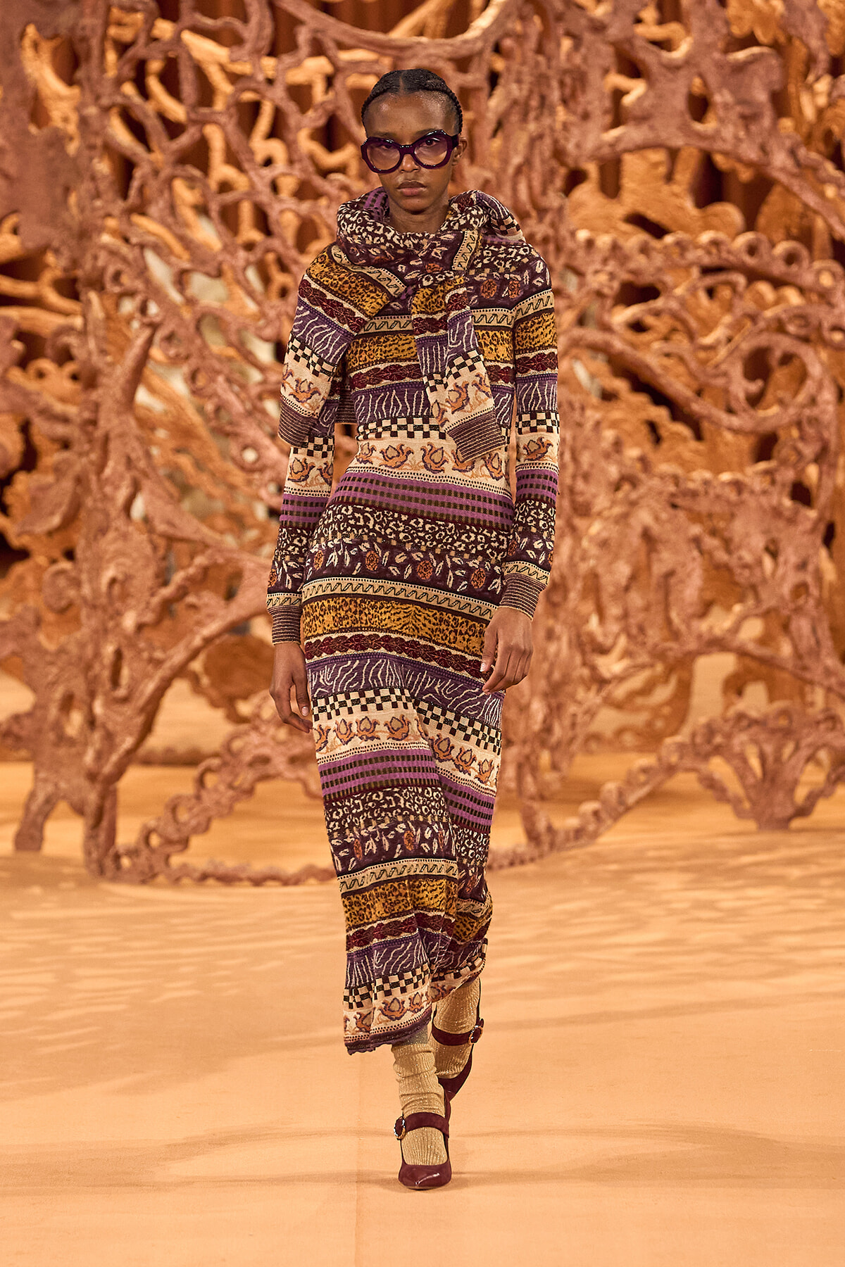 Model walking a runway in a multicolored patterned knit dress and scarf, wearing dark sunglasses, with a brown sculptural backdrop behind her.