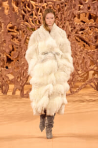 Female model walks a fashion runway in an oversized white fluffy fur coat with a belt, paired with gray knee-high boots, against a brown sculptural backdrop.