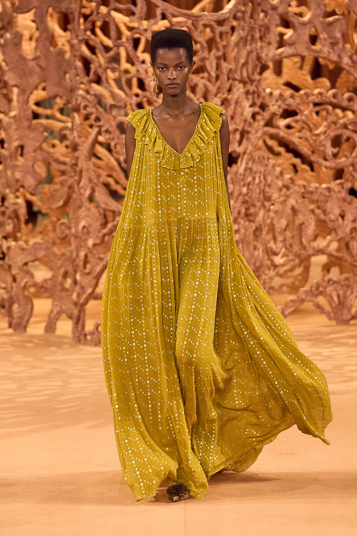 Model walking the runway in a mustard-yellow, ruffled V-neck maxi dress with white speckles.