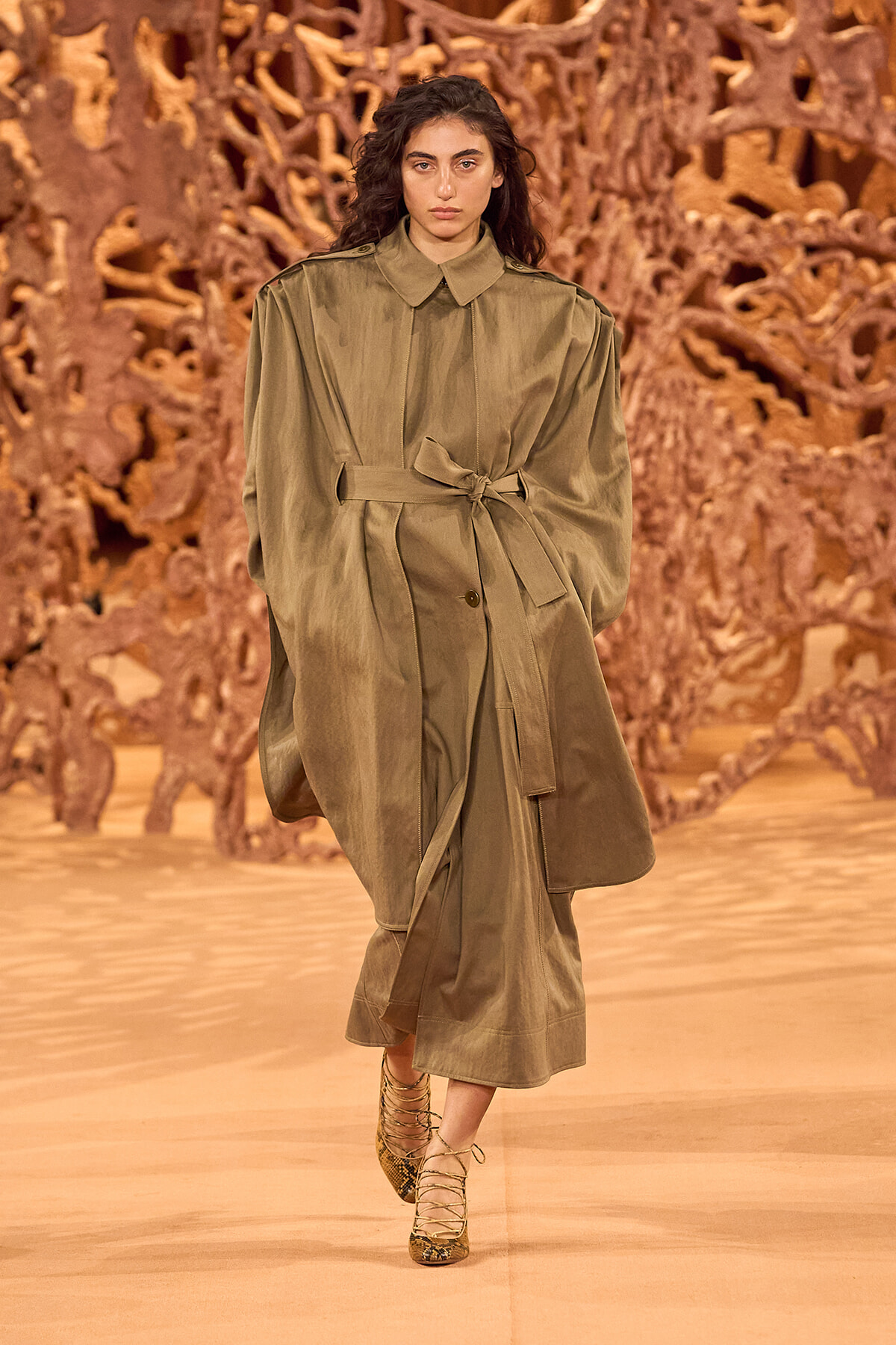 Model walks the runway in an olive trench coat with a tied belt, matching pants, and snakeskin heels; an orange sculptural backdrop surrounds the scene.