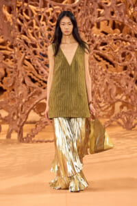 Model walking runway in olive green sleeveless V-neck top with white pinstripes and metallic gold pleated pants, holding a gold bag against a sculptural brown backdrop.