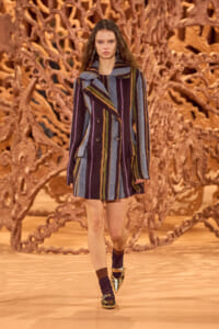 Model on a runway wearing a long multicolored striped coat with a wide collar and double-breasted buttons; brown coral-like sculpture backdrop behind her.