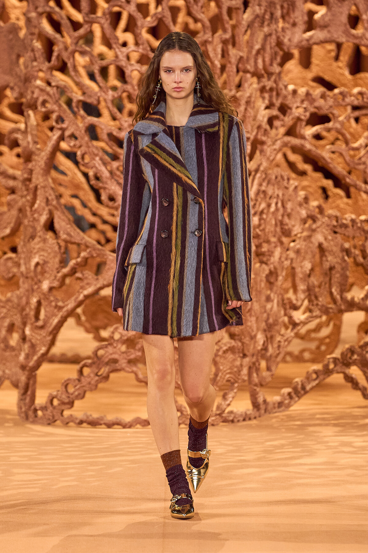 Model on a runway wearing a long multicolored striped coat with a wide collar and double-breasted buttons; brown coral-like sculpture backdrop behind her.