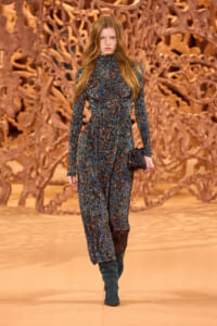 Model walking a runway in a long-sleeve teal-blue abstract-print dress with a high neckline, carrying a small black clutch.