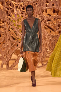 Model walks a fashion runway wearing a dark green leather sleeveless wrap dress with a waist tie, paired with brown leather pants and a two-tone bag; textured sculpted backdrop behind.