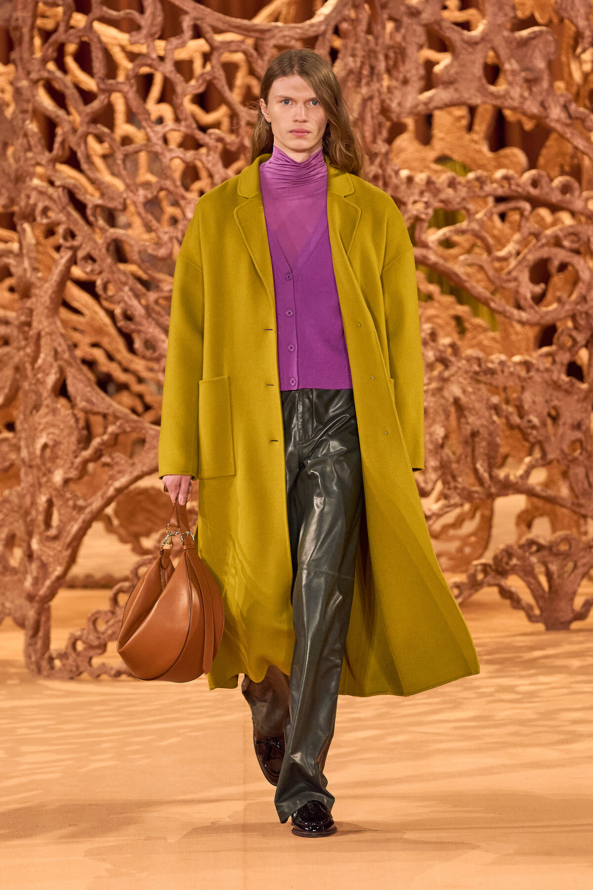 Model walking runway in a mustard yellow long coat over a purple turtleneck and dark green leather pants, carrying a brown handbag against a sculptural wooden backdrop.