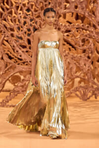 Model walks the runway in a gold metallic pleated gown with thin straps, against an ornate brown sculptural backdrop.