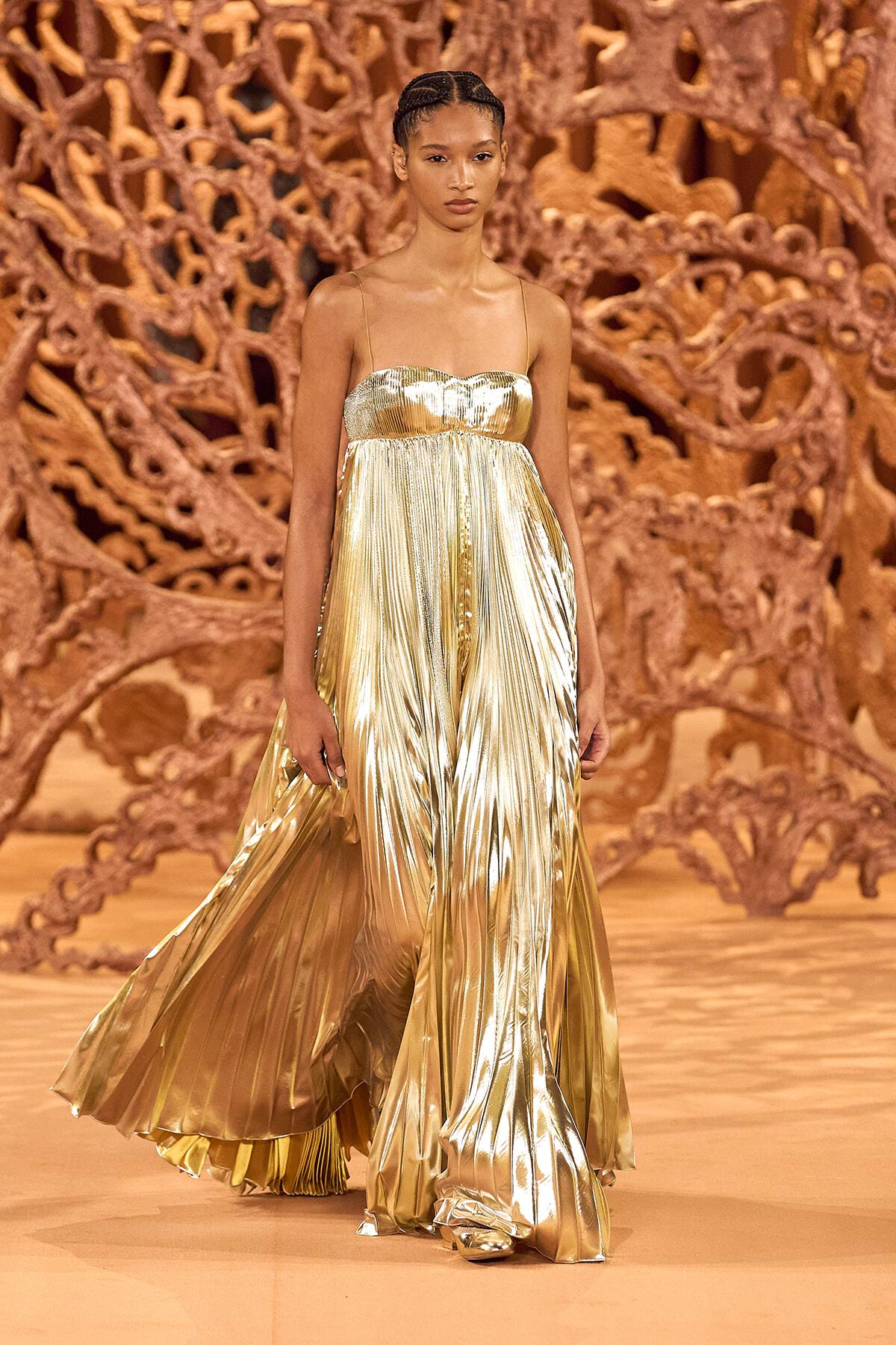 Model walks the runway in a gold metallic pleated gown with thin straps, against an ornate brown sculptural backdrop.