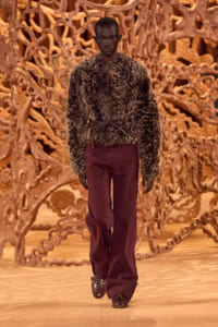 Model wearing a thick fur jacket and burgundy pants on a fashion runway with a sculptural wooden backdrop.