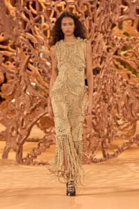 Model walks the runway in a beige intricate crochet dress with fringe, against a sculptural brown backdrop.