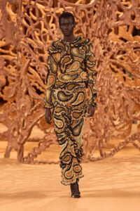 Model walks a runway in a bold beige, black, and gold paisley-print outfit with ruffled edges; sculptural brown backdrop behind.