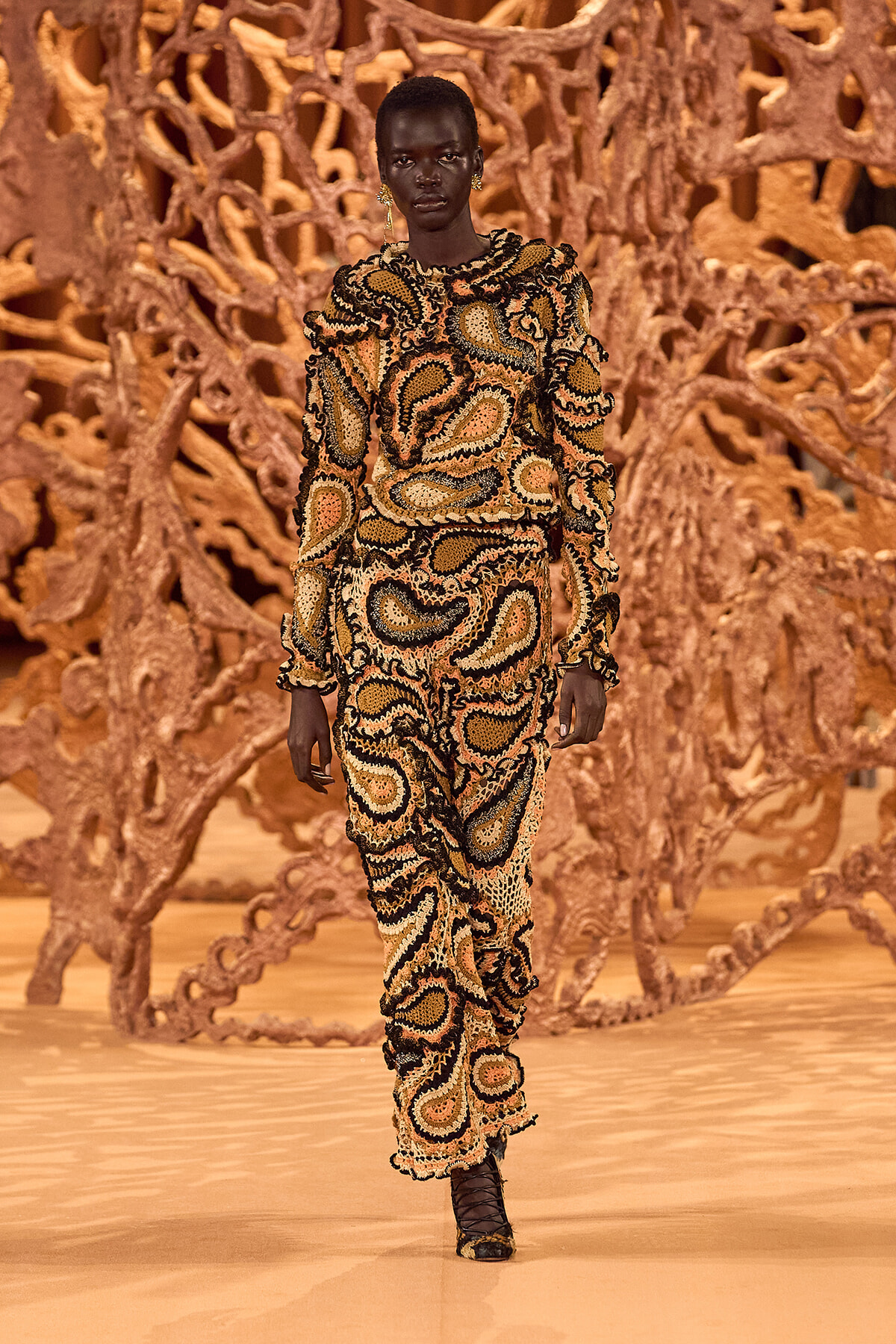 Model walks a runway in a bold beige, black, and gold paisley-print outfit with ruffled edges; sculptural brown backdrop behind.