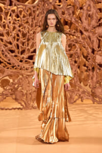 Model on a fashion show runway wearing a two-tone metallic, pleated gown in gold and silver with an intricate sculptural backdrop behind her.