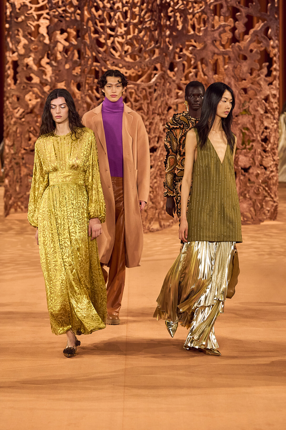Models walk a runway in gold, bronze, and olive-toned outfits against an intricate, brown sculptural backdrop.