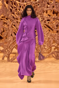 Model walks a runway in a purple dress with dramatic ruffles and oversized sleeves, holding a small matching handbag.