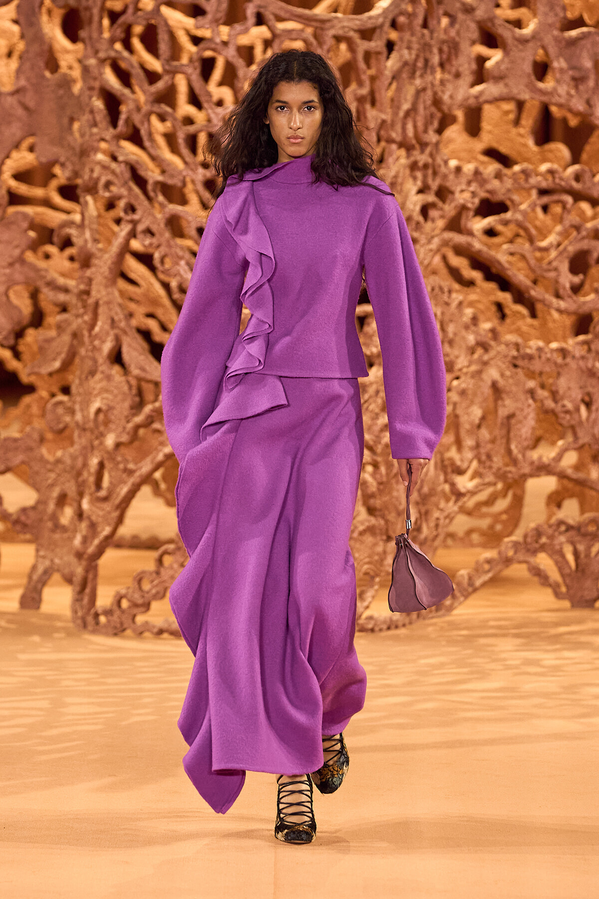 Model walks a runway in a purple dress with dramatic ruffles and oversized sleeves, holding a small matching handbag.