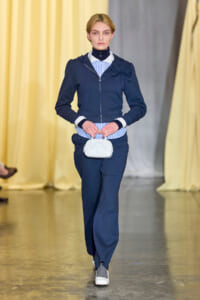Runway model in a navy zip-front jacket with rosette shoulder detail, striped shirt, navy pants, and a small white handbag.