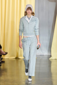 Model walking a fashion show runway in a light gray zip-up hoodie and joggers with rose appliqués, paired with a pale knit bonnet and white sneakers.