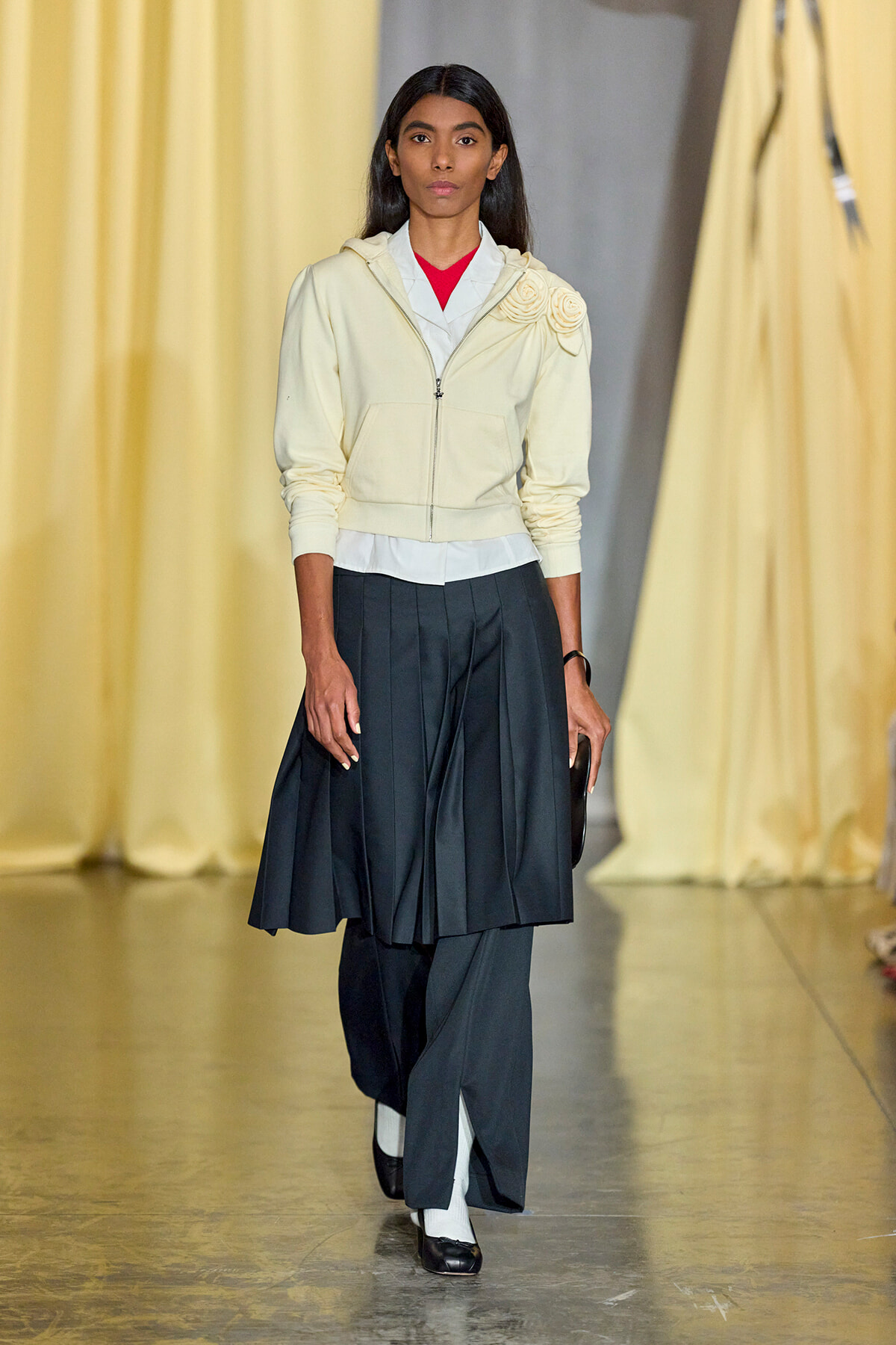Model walking the runway in a cream zip hoodie with floral shoulder embellishments, layered white shirt, red top, and dark pleated skirt with flats.