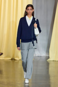Model walking a runway in a navy V-neck sweater over a white blouse, layered gray wide-leg pants, white sneakers, and a white handbag with a dark flower brooch.