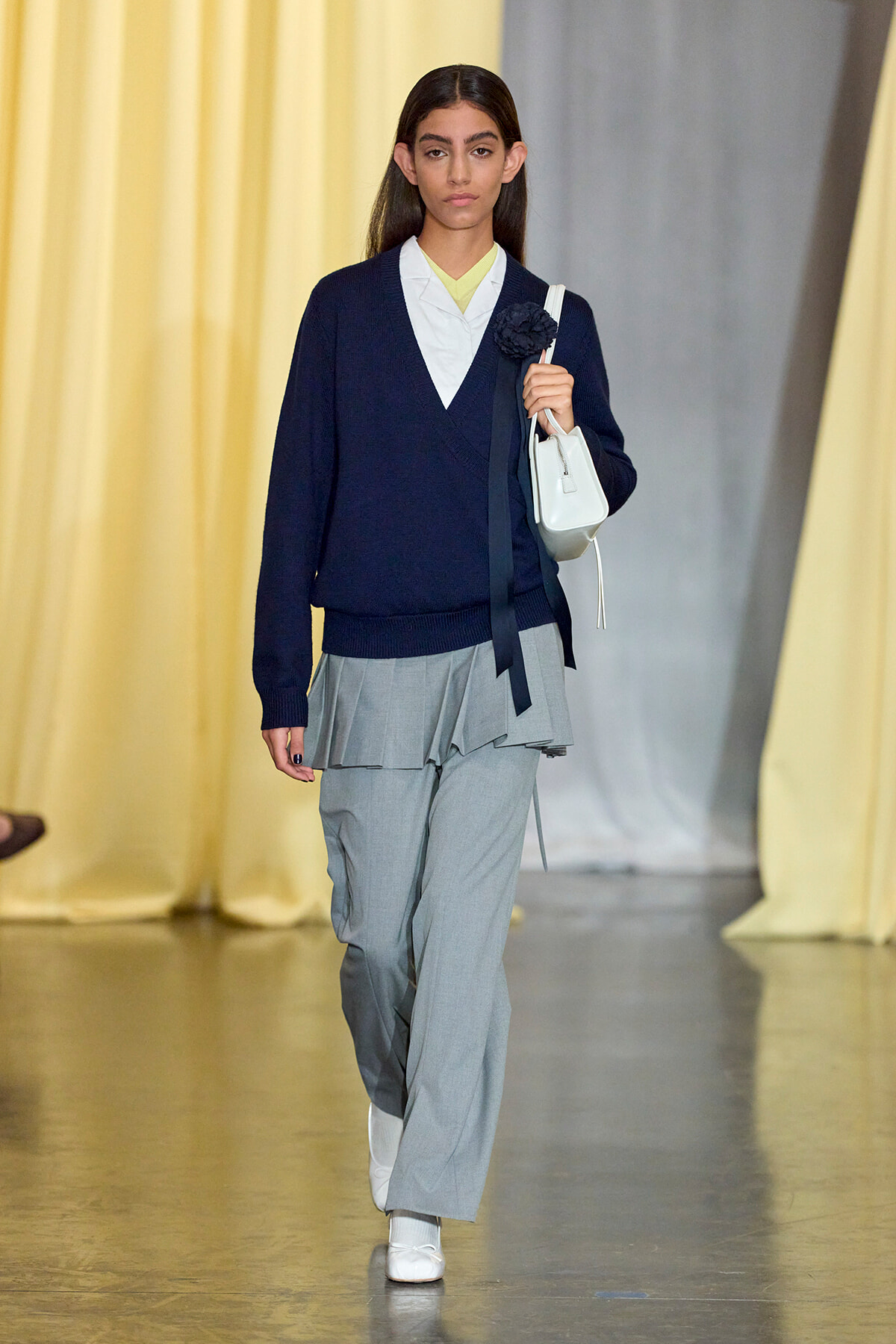 Model walking a runway in a navy V-neck sweater over a white blouse, layered gray wide-leg pants, white sneakers, and a white handbag with a dark flower brooch.