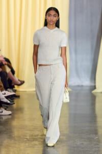 Model walking a fashion runway in a white knit top and pale gray wide-leg pants, carrying a small white handbag.