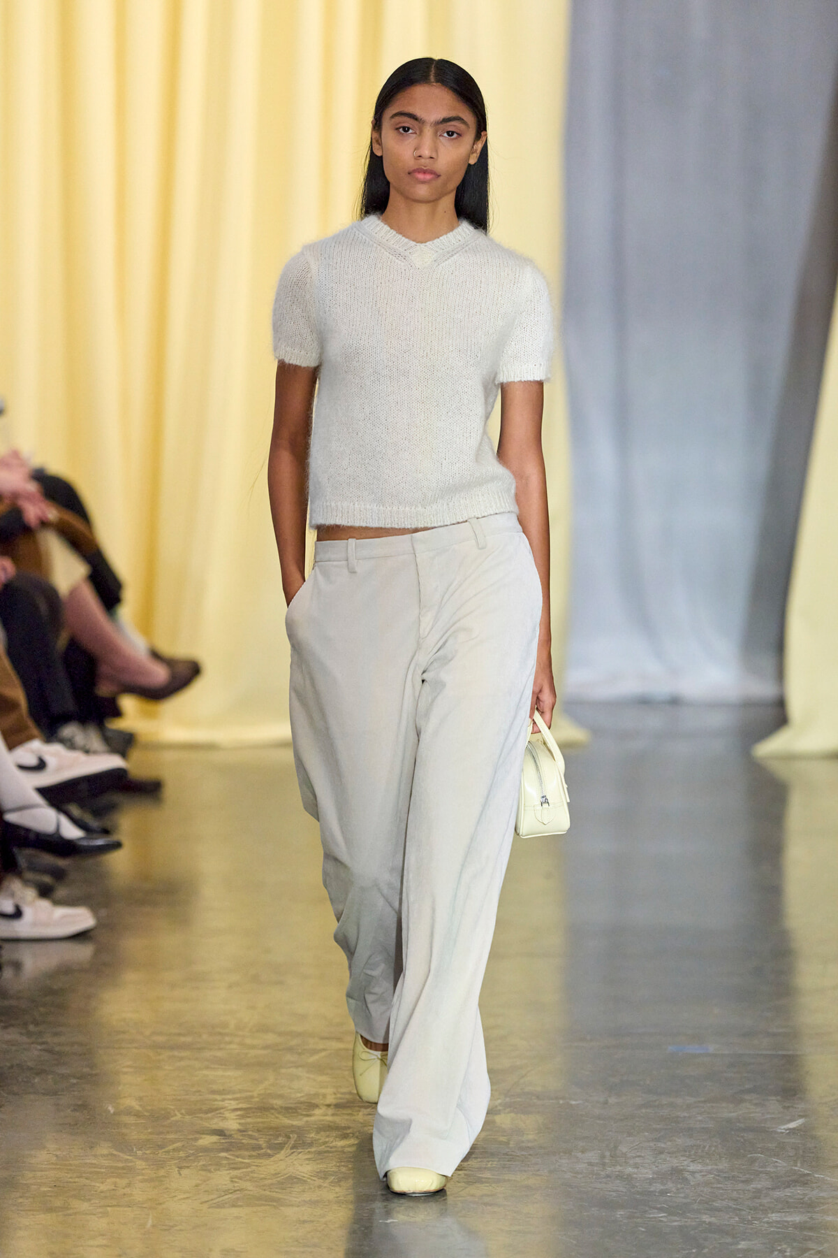 Model walking a fashion runway in a white knit top and pale gray wide-leg pants, carrying a small white handbag.