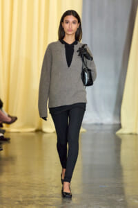 Model walking a fashion runway in a gray knit sweater over black top, black leggings, gloves, and a small black handbag.