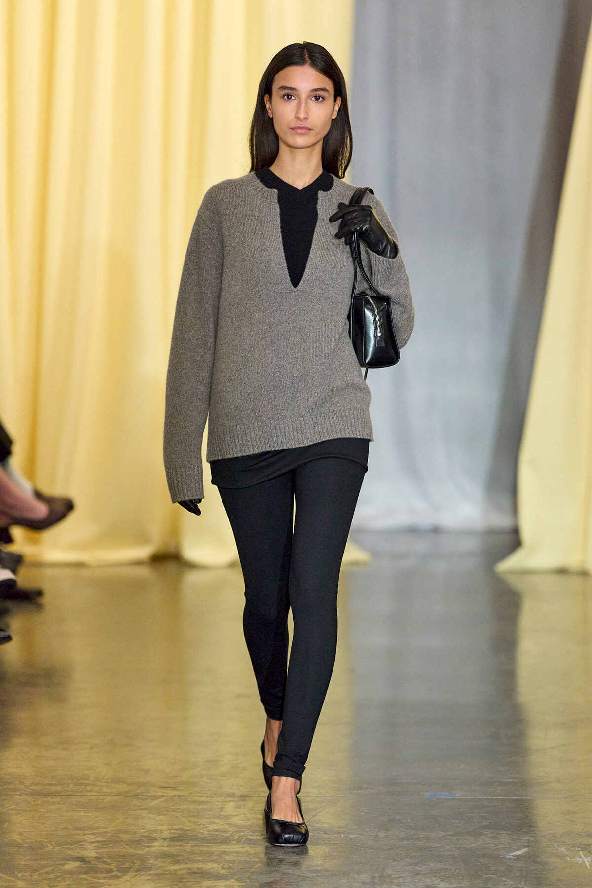 Model walking a fashion runway in a gray knit sweater over black top, black leggings, gloves, and a small black handbag.