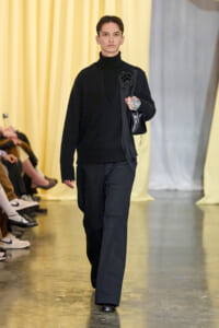 Model walking the runway in an all-black outfit: turtleneck sweater, wide-leg pants, and a small black shoulder bag, with a flower brooch and visible tattoo on the hand against a yellow backdrop.