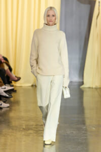 Model walking a fashion show in a cream knit turtleneck sweater and light wide-leg pants, carrying a small white purse.