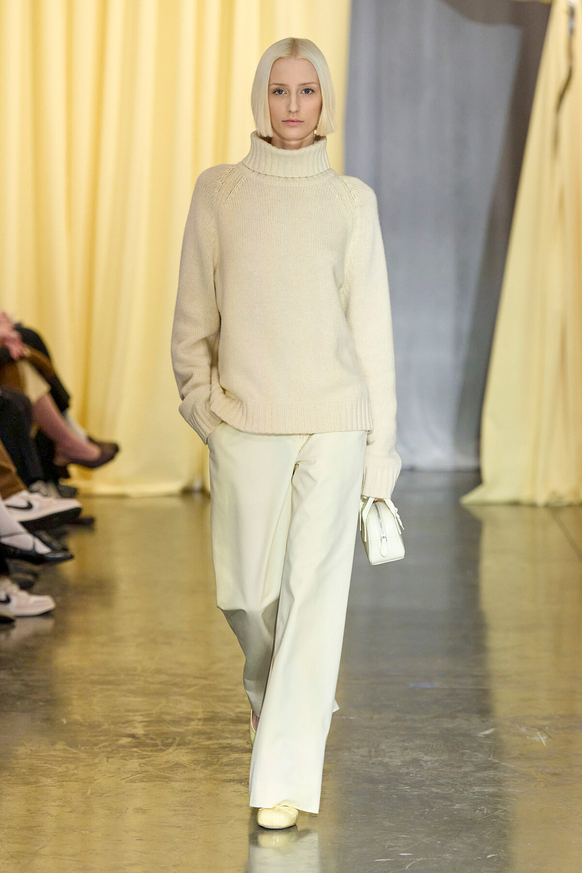 Model walking a fashion show in a cream knit turtleneck sweater and light wide-leg pants, carrying a small white purse.