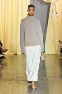 Model walking a fashion runway in a gray knit turtleneck sweater and white trousers, holding a small beige handbag.