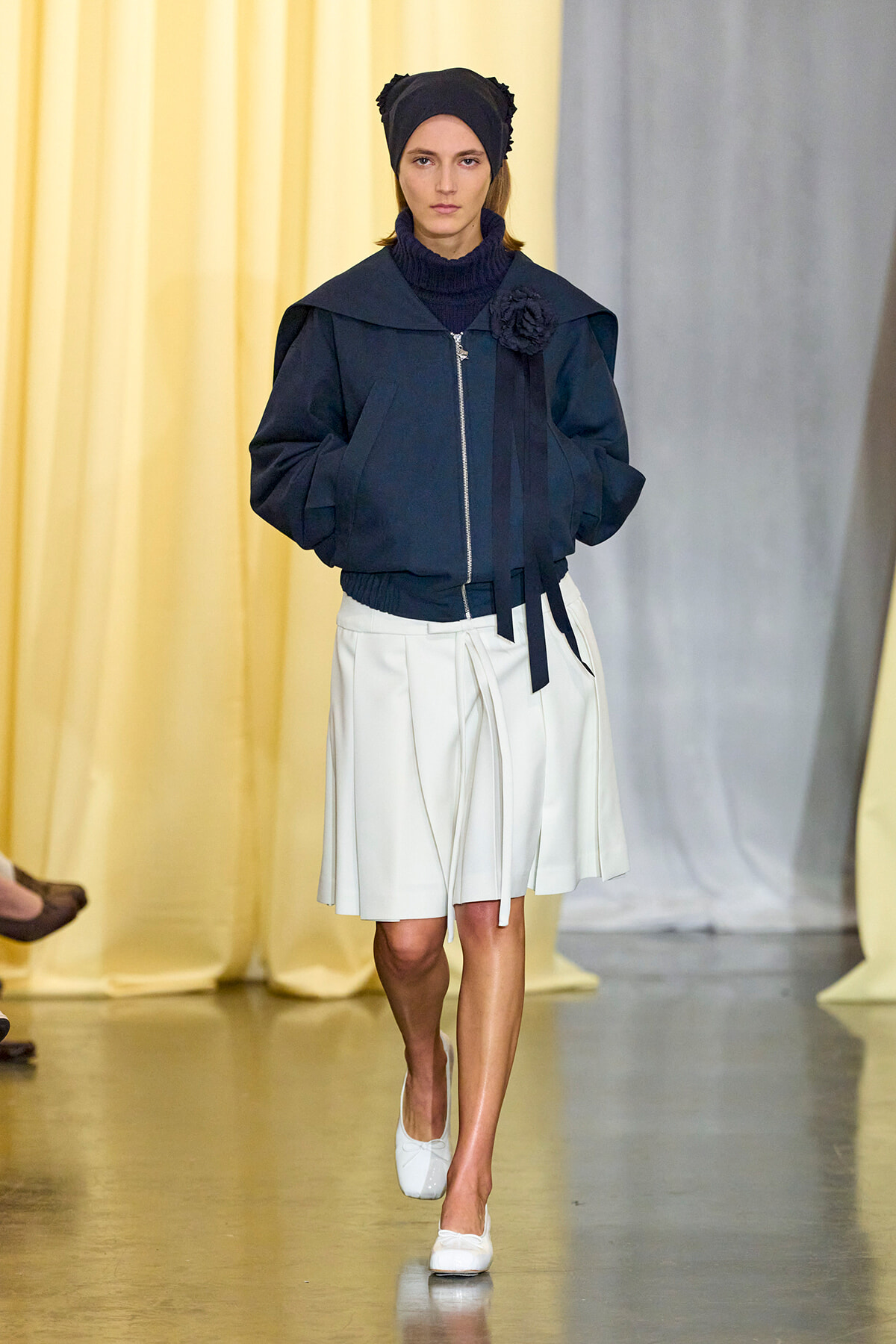 Model on runway wearing a dark oversized navy jacket with a flower brooch, white pleated skirt, and white flats.