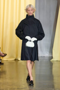 Model walking the runway in a black high-neck coat, white gloves, and a small white handbag.