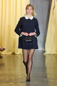 Model walks the runway in a navy dress with a white Peter Pan collar, clutching a small black handbag.