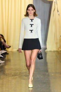 Model walks the runway in a pale blue/cream top with three black ribbon closures and a short black skirt, carrying a small black handbag.