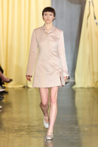 Model walks the runway in a pale pink tailored blazer dress, holding a metallic clutch with a soft yellow curtain backdrop.