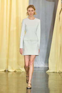 Model walking runway in a light gray dress with a belt, neutral-toned shoes, against pale yellow curtains.