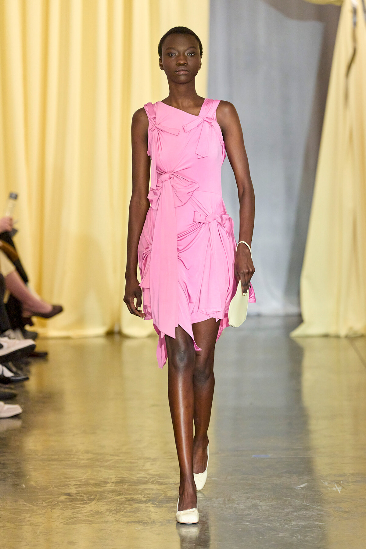 Model walking a fashion runway in a pink draped dress with bow details, holding a small white clutch.