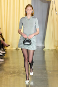 Model walks the runway in a pastel-blue zip-front top and fluffy mini skirt, holding a small black handbag.
