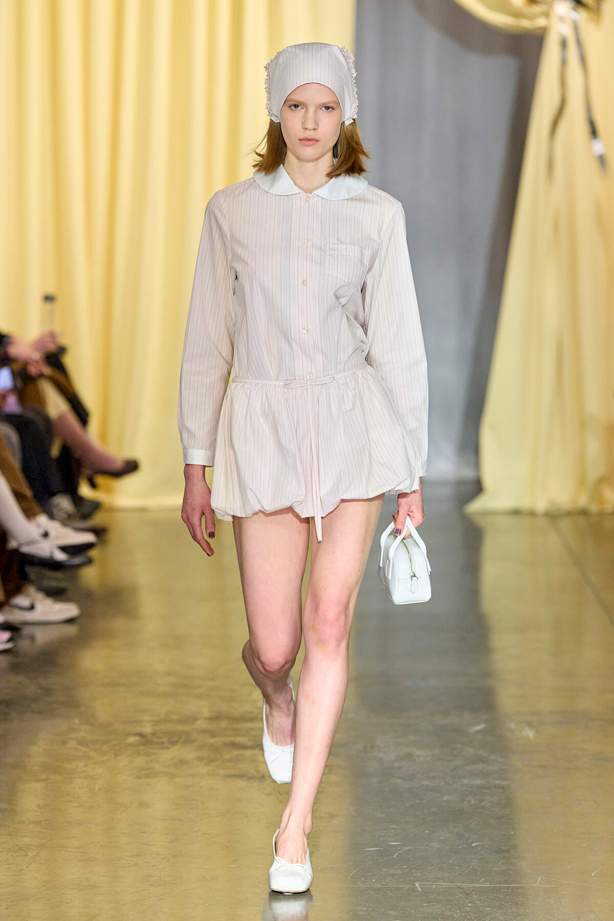 Model walking a fashion show in a pale striped shirt dress with a Peter Pan collar, white headscarf, and a small white handbag on the runway