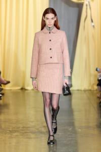 Model walking a fashion show in a pale pink tweed coat and matching skirt, buttoned front, carrying a small black handbag.
