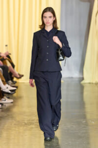 Model walking a fashion runway in a navy tailored blazer and matching wide-leg pants, carrying a small black handbag over her shoulder.