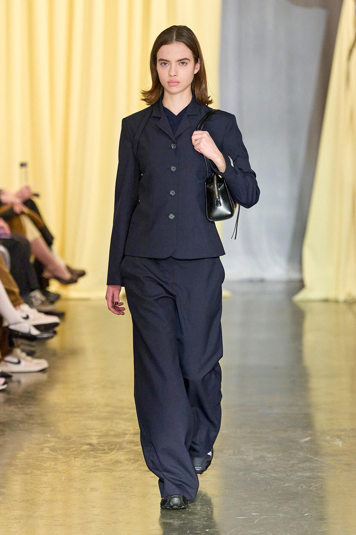 Model walking a fashion runway in a navy tailored blazer and matching wide-leg pants, carrying a small black handbag over her shoulder.