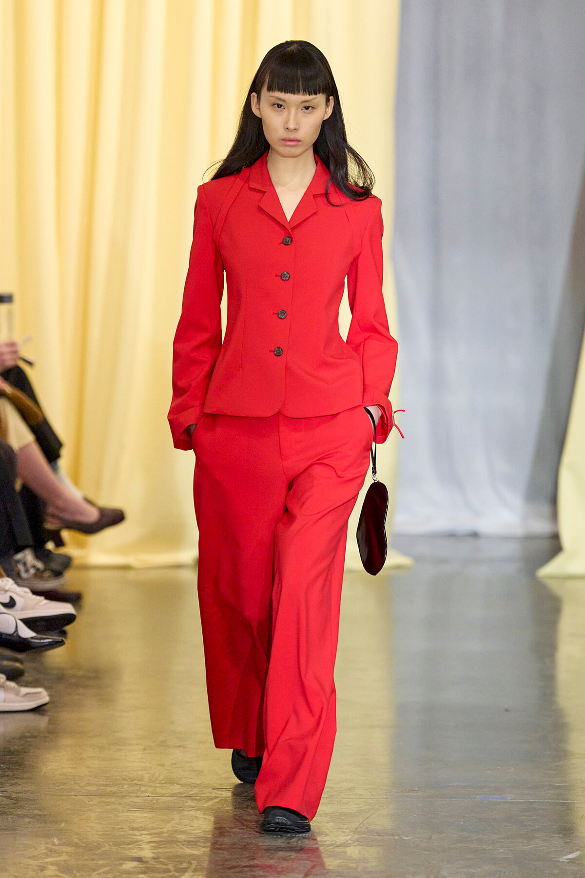 Model walking a fashion runway in a bright red tailored pantsuit with a matching blazer and wide-leg trousers, carrying a small black clutch.