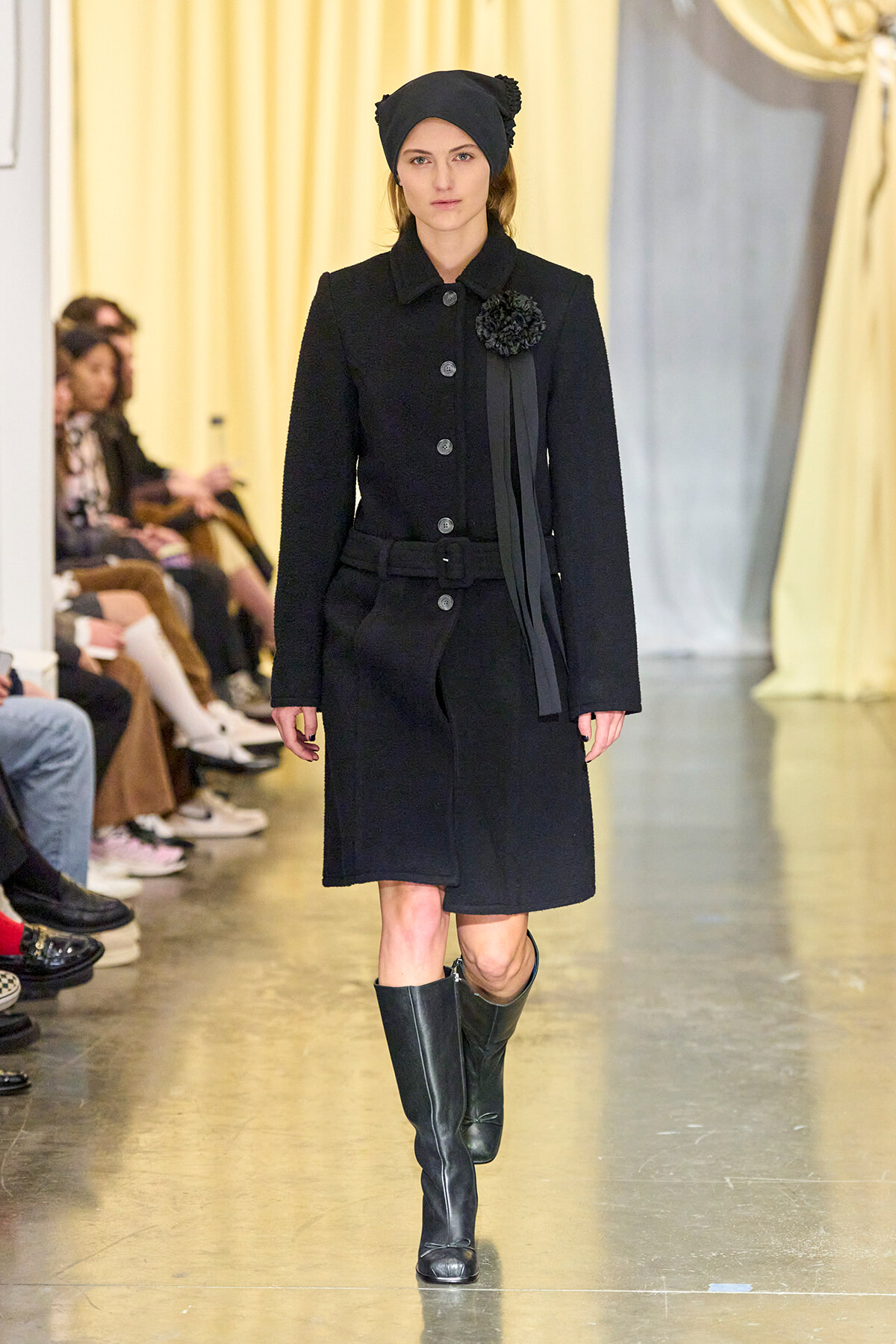 Female model walking a runway in a black knee-length coat with a belt, black hat, and tall boots.