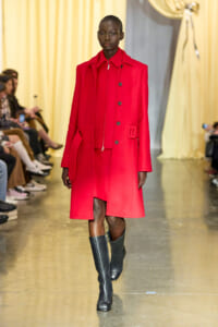 Model walking a runway in a bright red knee-length coat with a zip and buttons, paired with black knee-high boots.