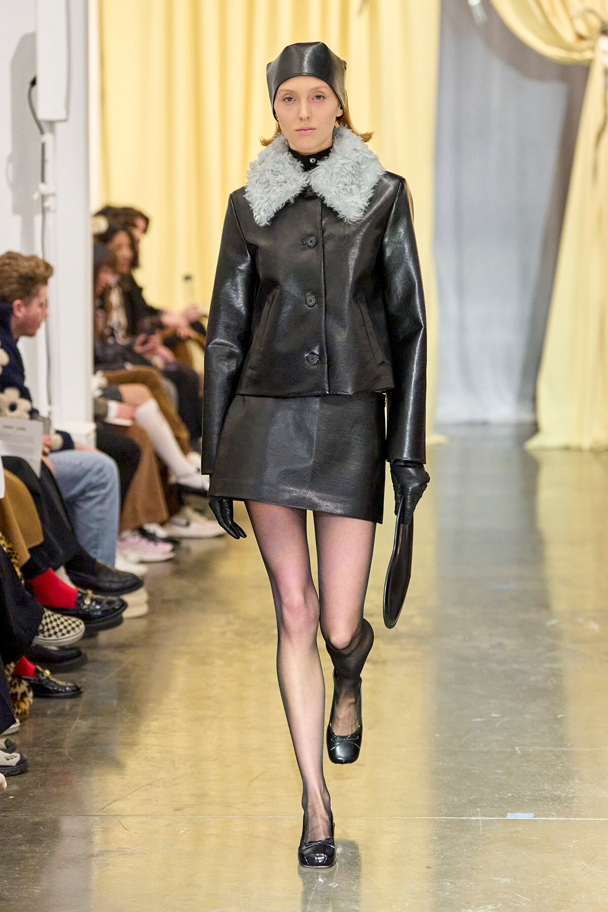 Model walking a fashion show wearing a black leather coat with a light gray fur collar, matching hat and gloves on a runway.