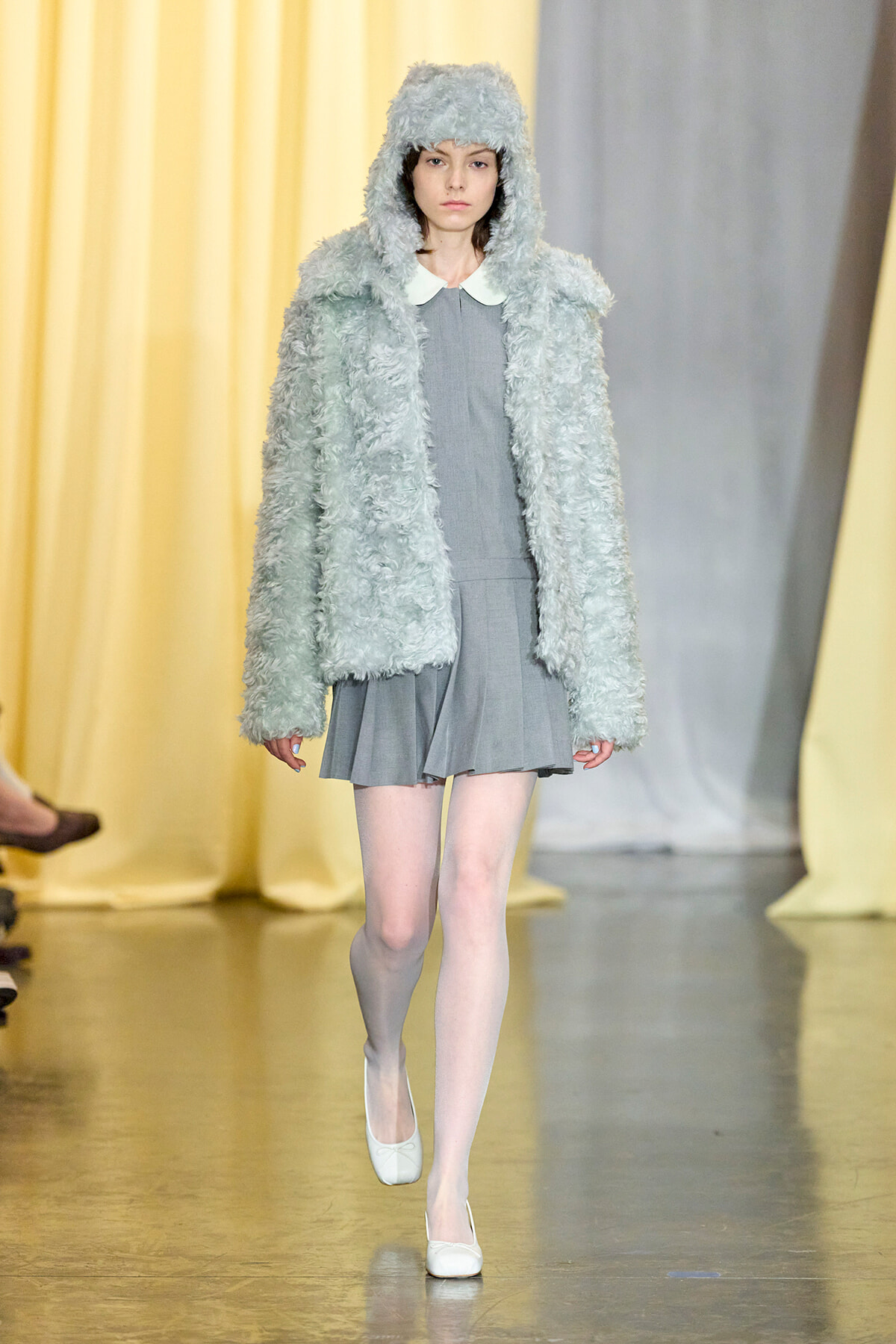 Model on a fashion show runway wearing a light gray fluffy hooded coat over a gray dress with a white collar, white tights and flats.